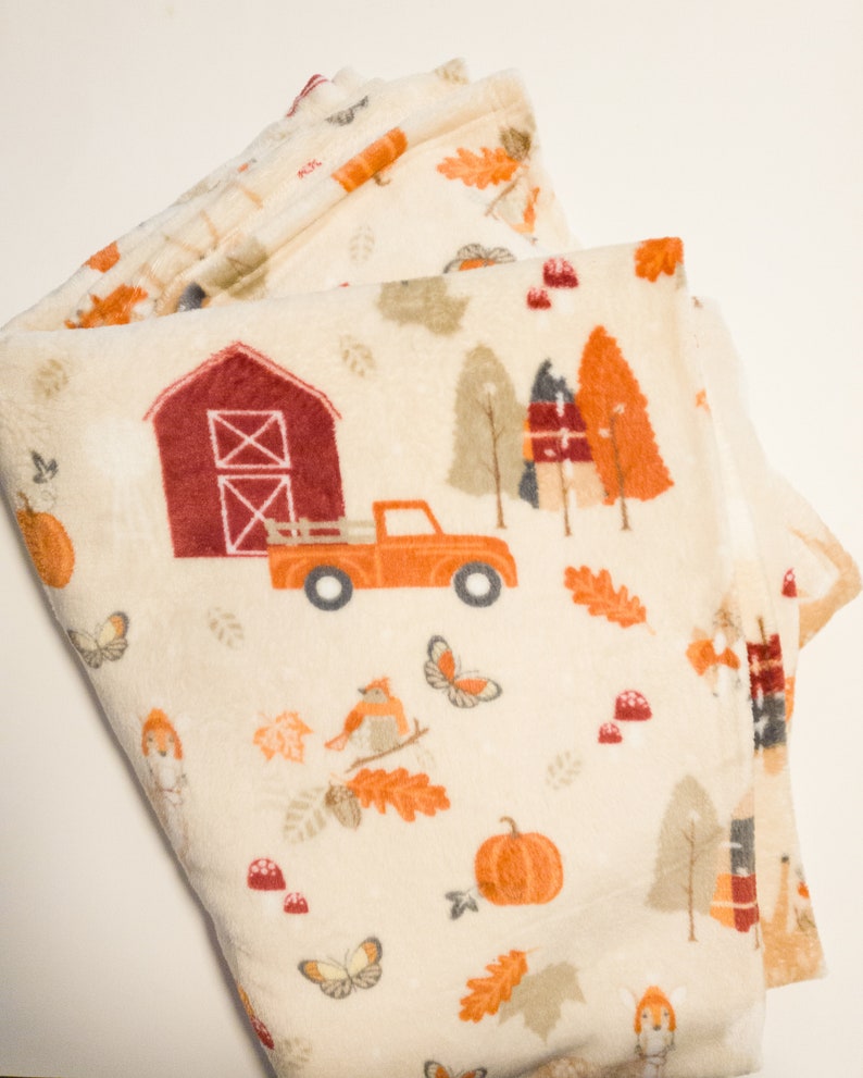 May include: A cream-colored fleece blanket with a fall-themed print. The print features a red barn, a red truck, pumpkins, leaves, and birds.