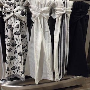 May include: Four kitchen towels hanging on a stainless steel oven. The towels include a black and white floral print, a white waffle weave, a white and gray striped design, and a solid black towel.