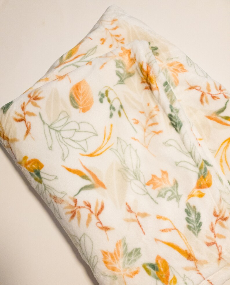 May include: A white plush throw blanket with a pattern of orange, yellow, and green leaves.