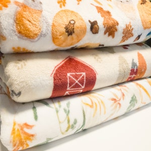 May include: Three white fleece blankets with different autumn-themed patterns. The top blanket has a pumpkin, leaves, and a small animal. The middle blanket has a red barn. The bottom blanket has a floral pattern with orange and green leaves.