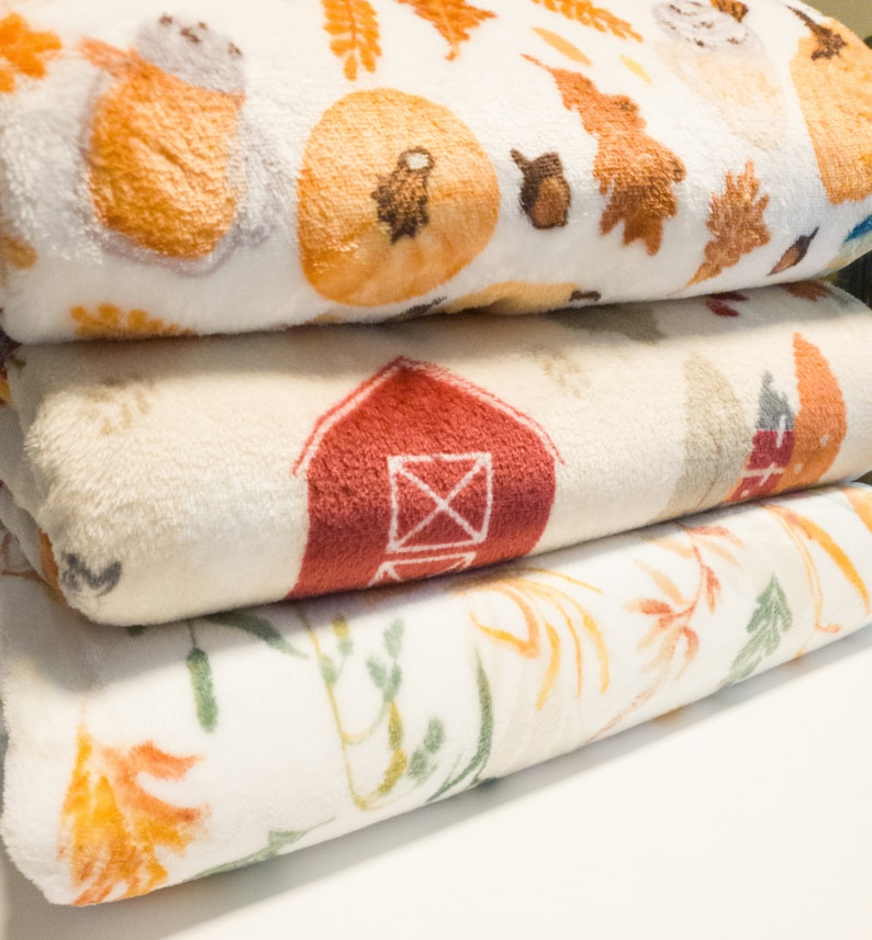 May include: Three white fleece blankets with different autumn-themed patterns. The top blanket has a pumpkin, leaves, and a small animal. The middle blanket has a red barn. The bottom blanket has a floral pattern with orange and green leaves.