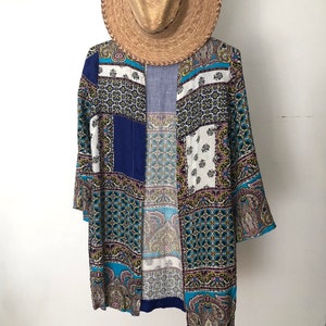 May include: A long, open-front kimono-style garment with a patchwork design in shades of blue, white, and teal, accented with paisley and geometric patterns. A straw hat hangs above the garment, suggesting a beach or resort theme.