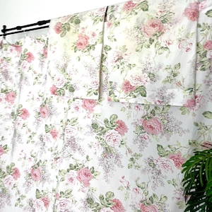 May include: A set of white curtains featuring a repeating floral pattern with pink roses, green leaves, and small purple flower clusters. The fabric is draped from a black curtain rod, displaying a soft, light-colored textile with botanical motifs.