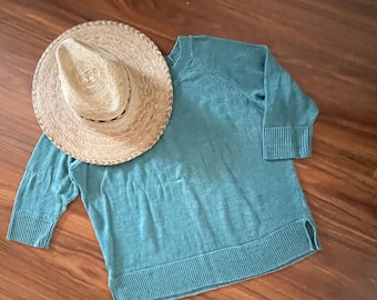 Puede incluir: Un suéter de color verde azulado con mangas tres cuartos y un sombrero de paja con ala tejida. El suéter tiene puños y dobladillo acanalados. El sombrero es de color marrón y redondo, con un ala ligeramente curvada. Los artículos están sobre una superficie de madera.