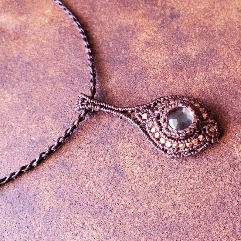 May include: A brown cord necklace with a brown macrame pendant featuring a clear gemstone.