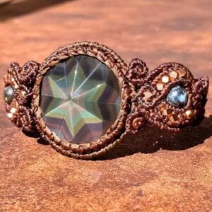 May include: A brown macrame bracelet with a large, iridescent, star-shaped crystal in the center. The bracelet is accented with smaller, blue crystals and gold beads.