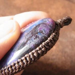 May include: A close-up of a dark purple and blue gemstone pendant with a brown cord. The gemstone has a swirling pattern and a smooth, polished surface.