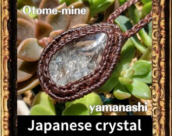 May include: A clear quartz crystal pendant with a brown leather cord. The pendant is set in a brown leather frame and has a natural, rough-cut shape. The text "Otome-mine yamanashi Japanese crystal" is visible in the image.