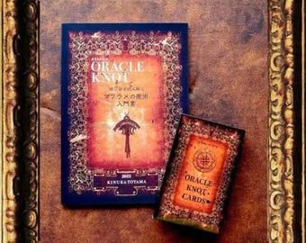 May include: A book titled "Oracle Knot" with a red, orange, and gold design. The book is about macrame magic and has a 2022 copyright date. A deck of cards titled "Oracle Knot Cards" is also shown.