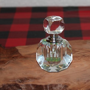 以下が含まれることがあります： 銀色のストッパーが付いた、透明なファセットクリスタル香水瓶が2つ、木製の表面に置かれています。ボトルは幾何学的な形状をしており、光を反射します。背景には赤と黒のチェック柄があります。