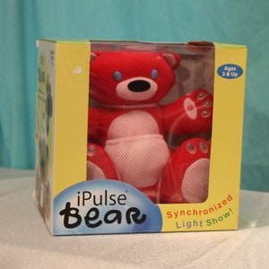 May include: A red iPulse Bear toy with a mesh texture, blue eyes, and a black nose, packaged in a yellow and blue cardboard box. The box features the text iPulse Bear and Synchronized Light Show, with a clear plastic window displaying the bear.