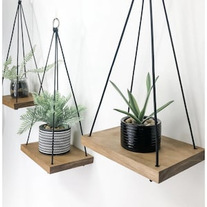 May include: Three wooden shelves with black rope hanging from the ceiling. Each shelf has a potted plant on it. The shelves are made of natural wood and the ropes are black.
