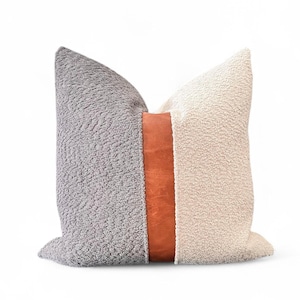 May include: A decorative pillow with a textured design. The pillow features a gray section on the left, a tan section on the right, and a brown leather strip down the center. The pillow is a square shape.