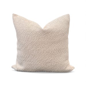 May include: A square, off-white decorative pillow with a textured, nubby surface. The pillow has a soft, inviting appearance and is likely made of a plush fabric. The pillow is a neutral color, making it suitable for various interior design styles.