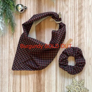 May include: A red and black plaid bandana and scrunchie set. The bandana is folded into a triangle shape and the scrunchie is round. The set is on a wooden background with Christmas ornaments.