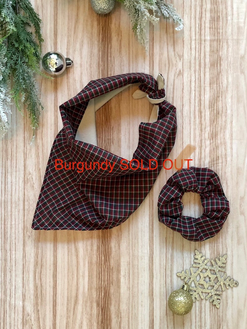 May include: A red and black plaid bandana and scrunchie set. The bandana is folded into a triangle shape and the scrunchie is round. The set is on a wooden background with Christmas ornaments.