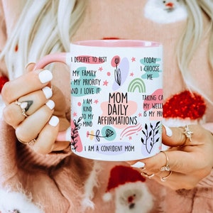 May include: A white ceramic mug with a pink rim and handle. The mug has a colorful design with the text "Mom Daily Affirmations" and positive affirmations for moms.