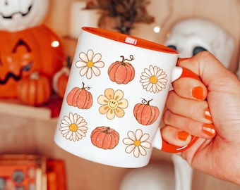 Op de afbeelding: Een witte keramische mok met een oranje rand, met een patroon van oranje pompoenen en witte madeliefjes. De mok wordt vastgehouden door een hand met oranje nagellak.
