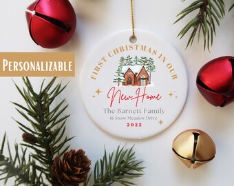 May include: White ceramic Christmas ornament with a red and brown illustration of a house with snow on the roof. The text "First Christmas in our New Home" is written in gold lettering around the house. The text "The Barnett Family, 16 Snow Meadow Drive, 2022" is written in black lettering below the house.