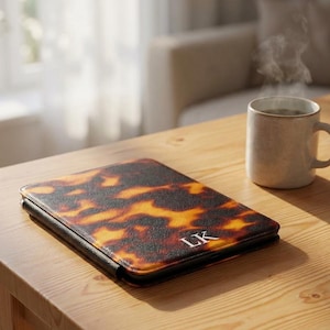 May include: A brown and yellow tortoiseshell-patterned tablet case with the initials "LK" in white. The case is on a wooden table next to a mug of coffee. The case has a black border.