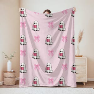 May include: A pink blanket patterned with white ghosts reading books and pink bows. The ghosts have black eyes and hold red books. The bows are a lighter pink. The blanket is held up by a person.