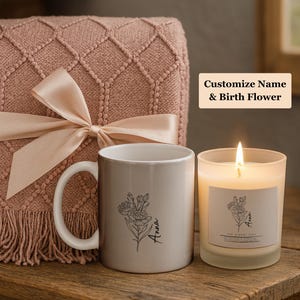 Op de afbeelding: Een beige keramische mok met een bloemenontwerp en de naam "Anna" staat naast een brandende kaars in een matglas. Een roze deken met een strik staat op de achtergrond. De tekst "Customize Name & Birth Flower" is zichtbaar.