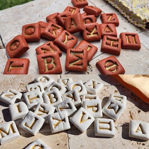 May include: Two sets of letter tiles, one set in red with gold lettering and one set in white with gold lettering. The tiles are arranged in a random pattern on a light brown surface.