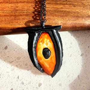 Puede incluir: Un collar con colgante negro con un centro naranja y amarillo llameante. El colgante tiene una forma estilizada única y cuelga de una cadena oscura. El centro del colgante tiene un efecto veteado arremolinado.