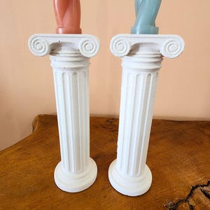 May include: Two white, fluted pillar candle holders with Ionic capitals. One holds a coral-colored candle, the other a pale blue candle. The candle holders are displayed on a wooden surface against a peach-colored background.