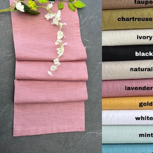 May include: A folded dusty rose linen table runner with a textured weave, decorated with a sprig of white flowers. A colour chart displays various linen colours, including taupe, chartreuse, ivory, black, natural, lavender, gold, white, and mint.