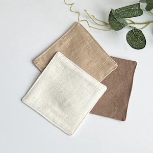May include: Three square fabric coasters in neutral tones: off-white, tan, and brown. The coasters are arranged in a layered pattern, showcasing their textured weave. A sprig of greenery is in the background.