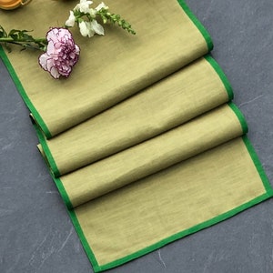 May include: A folded green and beige linen table runner with a green border. The runner is folded on a dark grey surface.