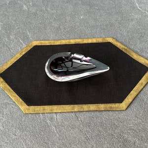 May include: A silver teardrop-shaped ashtray sits on a black hexagonal placemat with a gold border.