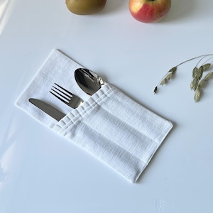 May include: A white linen cutlery holder with a fork, spoon, and knife. The silverware is stainless steel. A kiwi and a red apple are in the background. Dried wheat stalks are to the right.