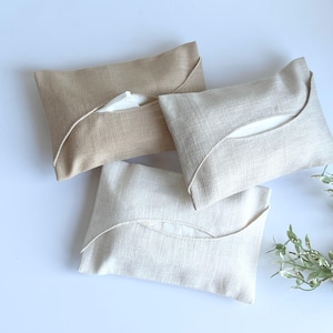 Puede incluir: Tres fundas de caja de pañuelos de lino en beige, blanco roto y marrón claro. Cada funda rectangular tiene una abertura curva para acceder a los pañuelos. Las fundas están dispuestas sobre una superficie blanca.