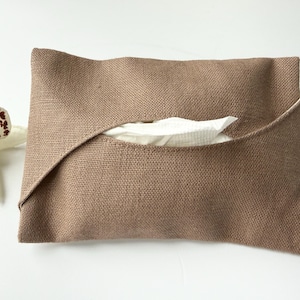 May include: A rectangular tissue holder made of a textured, light brown fabric with a folded overlapping design. A single white tissue is visible through the central opening. The holder has visible stitching along the edges and a woven appearance.