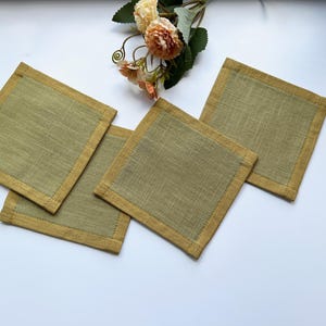 May include: A set of four square coasters featuring a sage green centre panel with a textured weave and a solid mustard yellow border. The coasters are arranged in a cluster with a small bouquet of artificial peach and cream-coloured flowers.