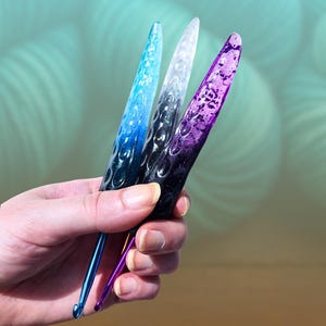 Puede incluir: Tres ganchillos con mangos de color degradado en azul, blanco y morado. Los mangos tienen un patrón texturizado, similar a burbujas. Los ganchillos se sostienen en una mano sobre un fondo verde claro borroso.