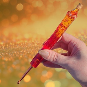 May include: A handcrafted crochet hook with a red and orange resin handle and a cork stopper. The hook has a copper-colored metal tip. The handle is held in a hand against a blurred, golden background.