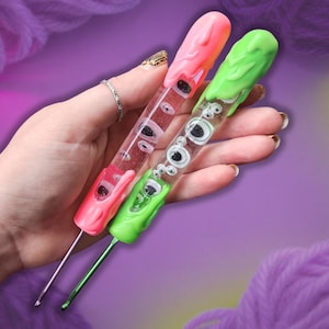 May include: Two crochet hooks with clear, cylindrical handles filled with small, multi-sized googly eyes. One hook features a bright pink, melted-wax style topper and base, while the other has a vibrant lime green, melted-wax style topper and base.