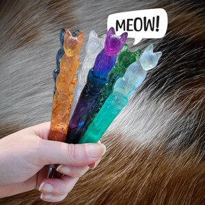 May include: A hand holds six cat-shaped resin wands in various colors: orange, clear, purple, green, and blue. A speech bubble above says "MEOW!" The wands are set against a blurred background of brown and gray fur.