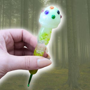 May include: A mushroom-shaped glass art piece with a light green stem and a white cap adorned with colorful dots. The stem has green and yellow accents. A green, teardrop-shaped detail is at the bottom.