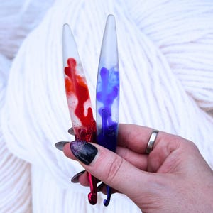 May include: Two crochet hooks with clear resin handles. One hook has red swirls, the other has blue swirls. The hooks are held in front of a background of white yarn.