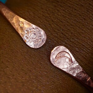 May include: A hammered copper bracelet with a sun and moon design. The bracelet is open and has a hammered finish.