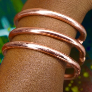 Op de afbeelding: Drie koperen armbanden gestapeld op de pols van een persoon. De armbanden zijn glad en glanzend, met een warme, roze gouden tint.