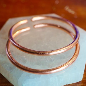 May include: Two copper bangles stacked on top of each other on a white surface.