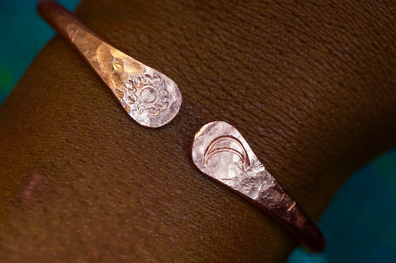 May include: A hammered copper bracelet with a sun and moon design. The bracelet is open and has a hammered finish.