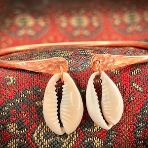 Op de afbeelding: Een koperen armband met twee witte kauri-schelpen eraan bevestigd. De armband is gehamerd en heeft een rustieke uitstraling.
