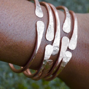 Puede incluir: Un conjunto de cinco pulseras de cobre martilladas con un acabado texturizado. Las pulseras est&aacute;n apiladas en la mu&ntilde;eca de una persona.