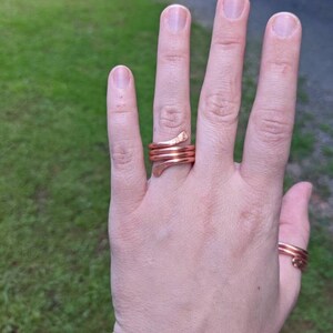 以下が含まれることがあります： 銅製のリング3つをつけた手のアップ。リングは厚みがあり、波打つような有機的なデザイン。人差し指につけています。